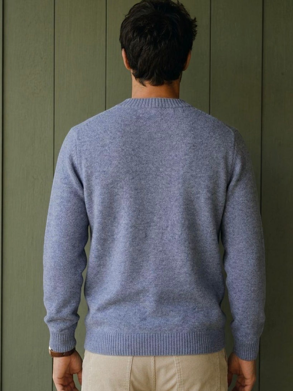 Back view of a man wearing a blue KZ DIFFUSION wool blend sweater with ribbed cuffs against a green wooden wall