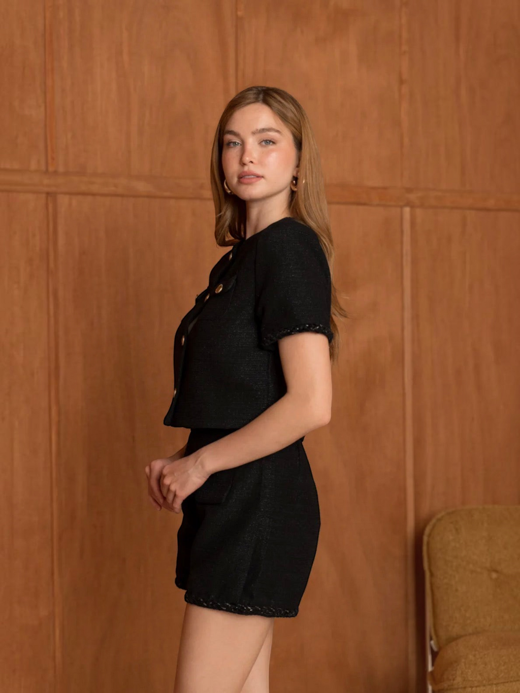 Top manches courtes noir BROCK en tissu texturé avec boutons dorés porté par une femme devant un mur en bois