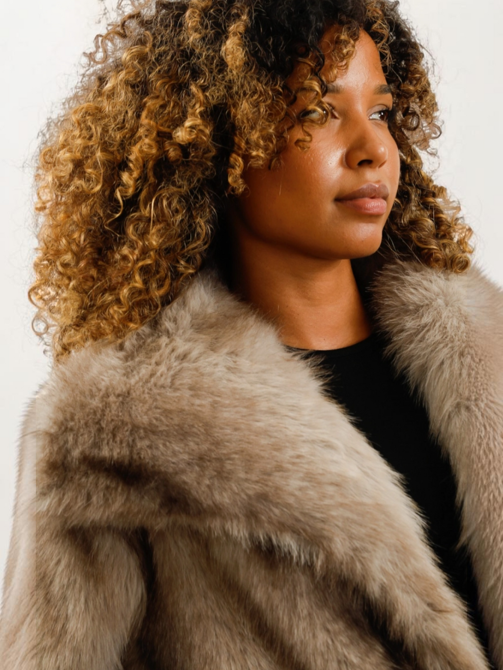 Femme aux cheveux bouclés portant un manteau oversize en fausse fourrure beige à col large de la marque BROCK
