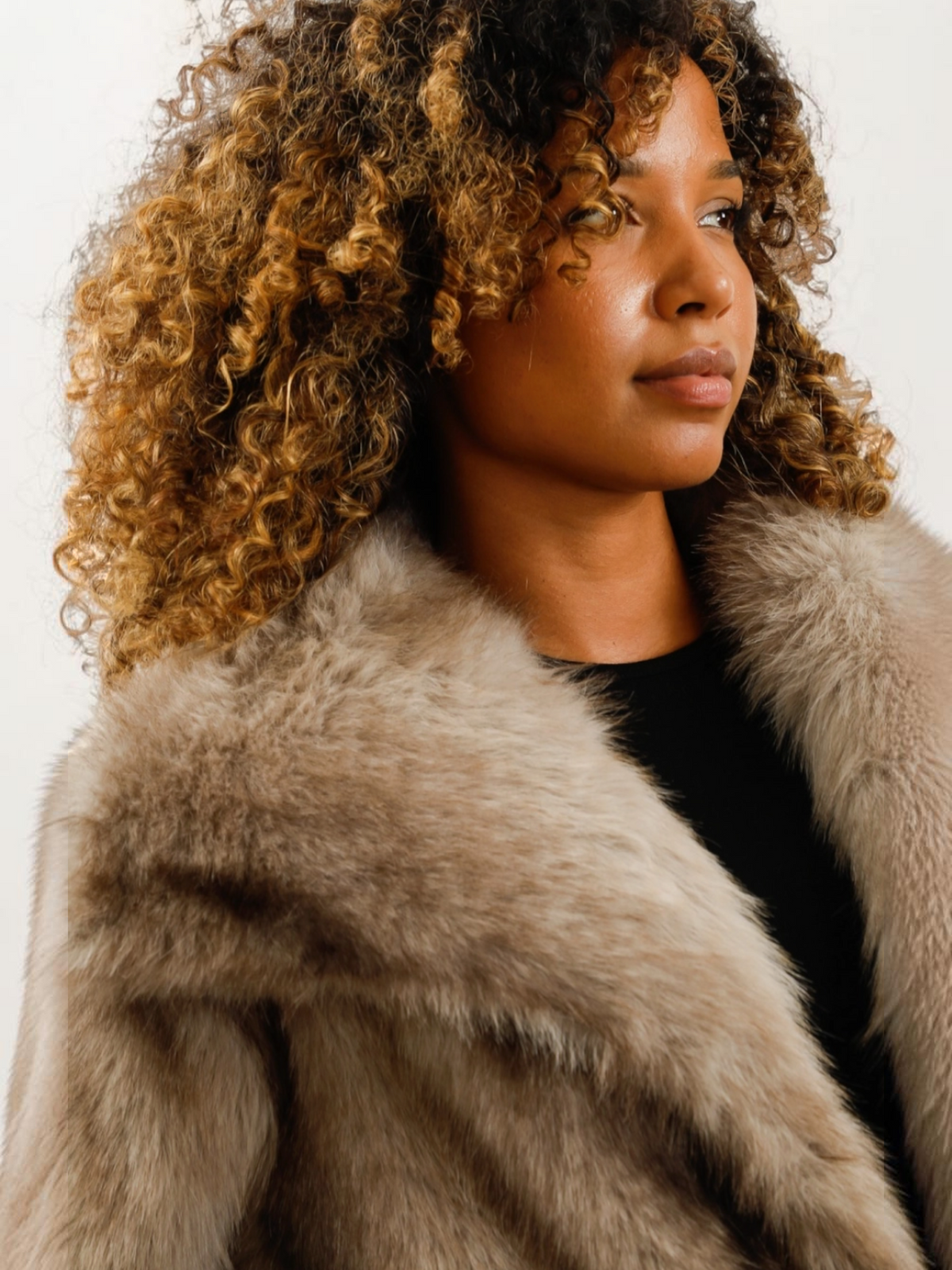 Femme aux cheveux bouclés portant un manteau oversize en fausse fourrure beige à col large de la marque BROCK