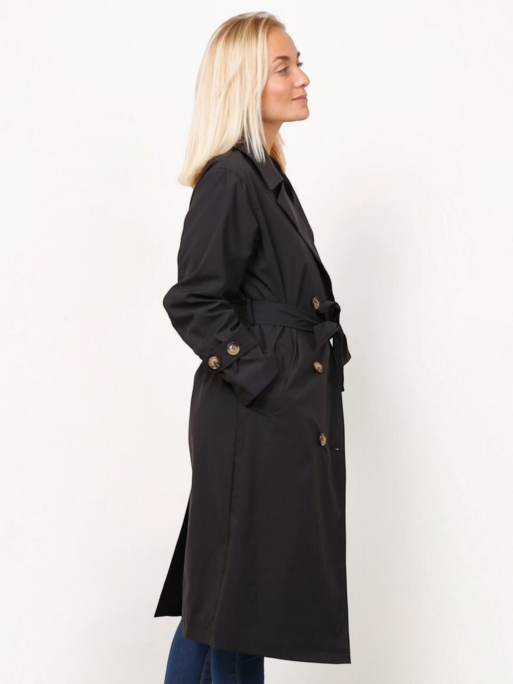 Femme blonde portant un trench long noir BROCK à double boutonnage avec ceinture et pattes de serrage aux poignets
