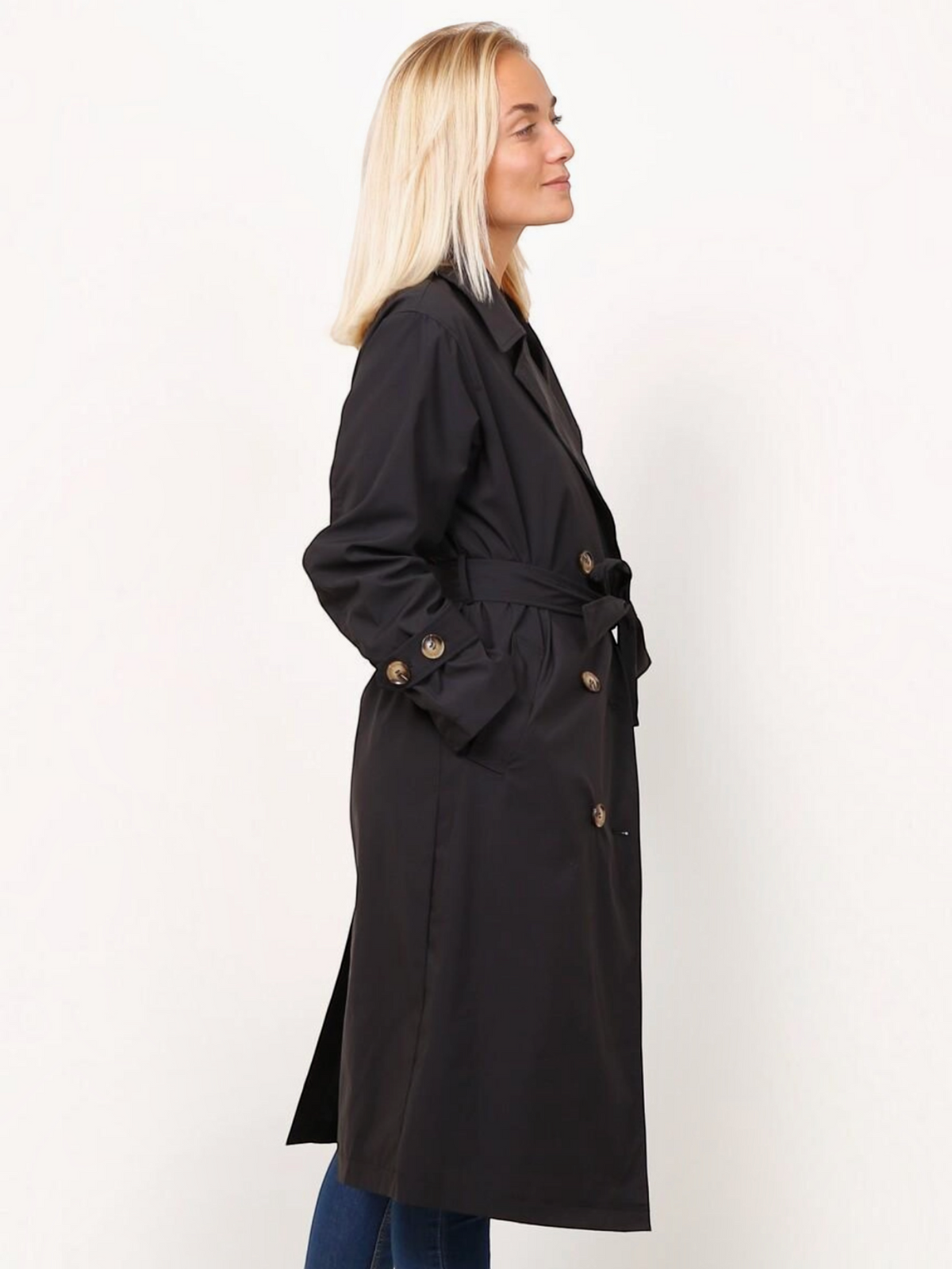 Femme blonde portant un trench long noir BROCK à double boutonnage avec ceinture et pattes de serrage aux poignets