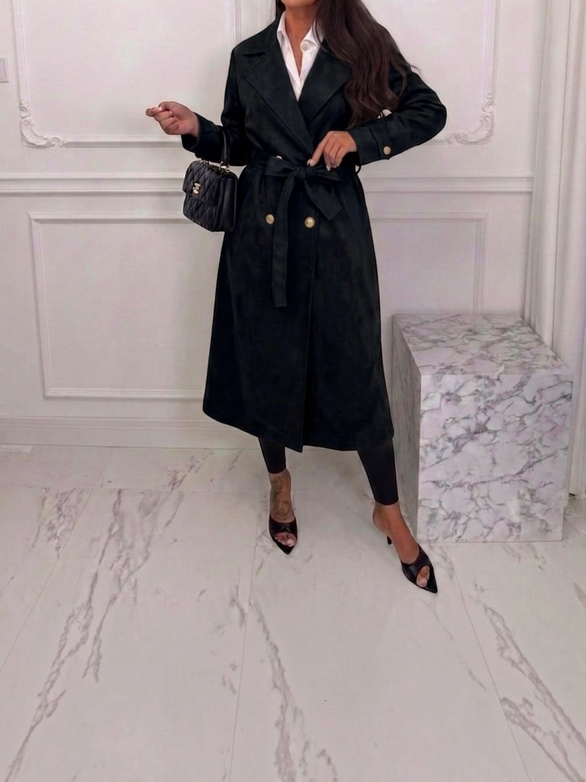 Trench long noir en suédine BROCK avec ceinture, porté par une femme avec chemise blanche et sac à main matelassé