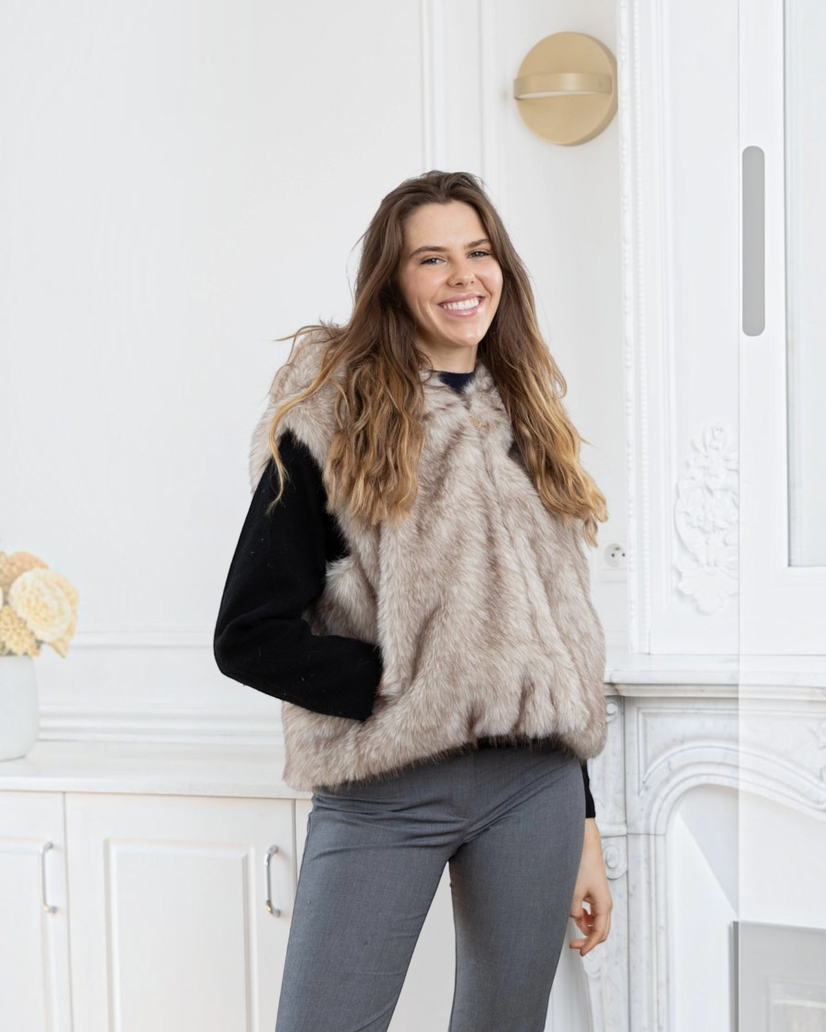 Woman wearing a BROCK beige two-tone faux fur hooded vest over a black sweater in a bright interior