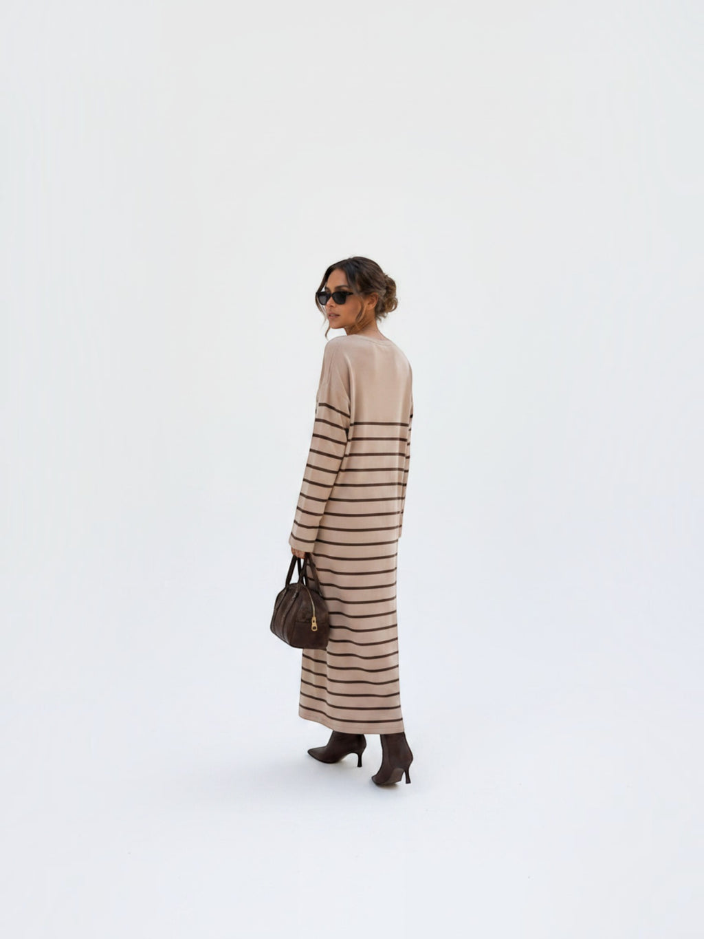 BROCK model wearing a beige maxi dress with brown stripes, V-neck, and long sleeves, styled with boots and a handbag.
