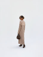 BROCK model wearing a beige maxi dress with brown stripes, V-neck, and long sleeves, styled with boots and a handbag.