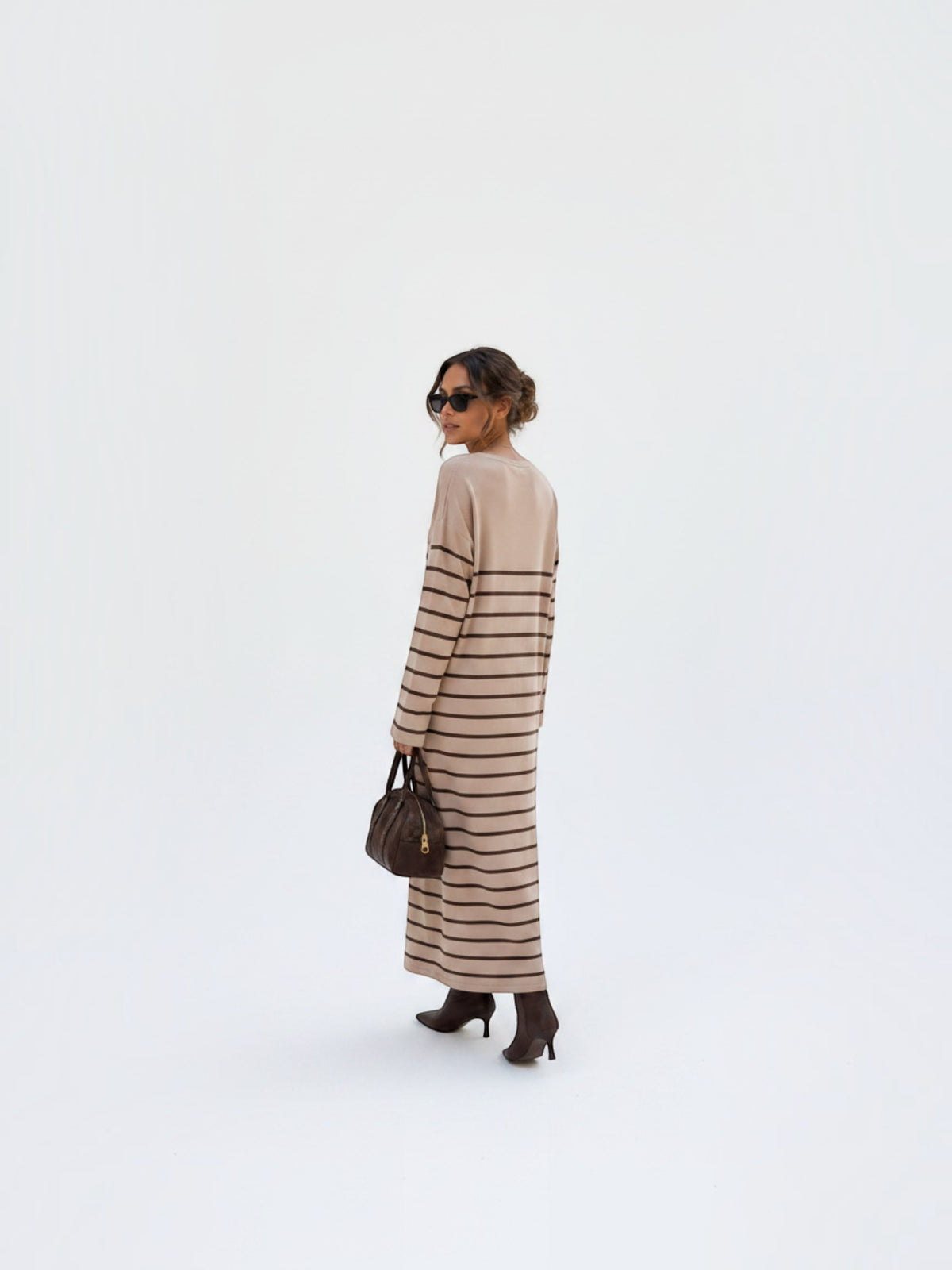 BROCK model wearing a beige maxi dress with brown stripes, V-neck, and long sleeves, styled with boots and a handbag.
