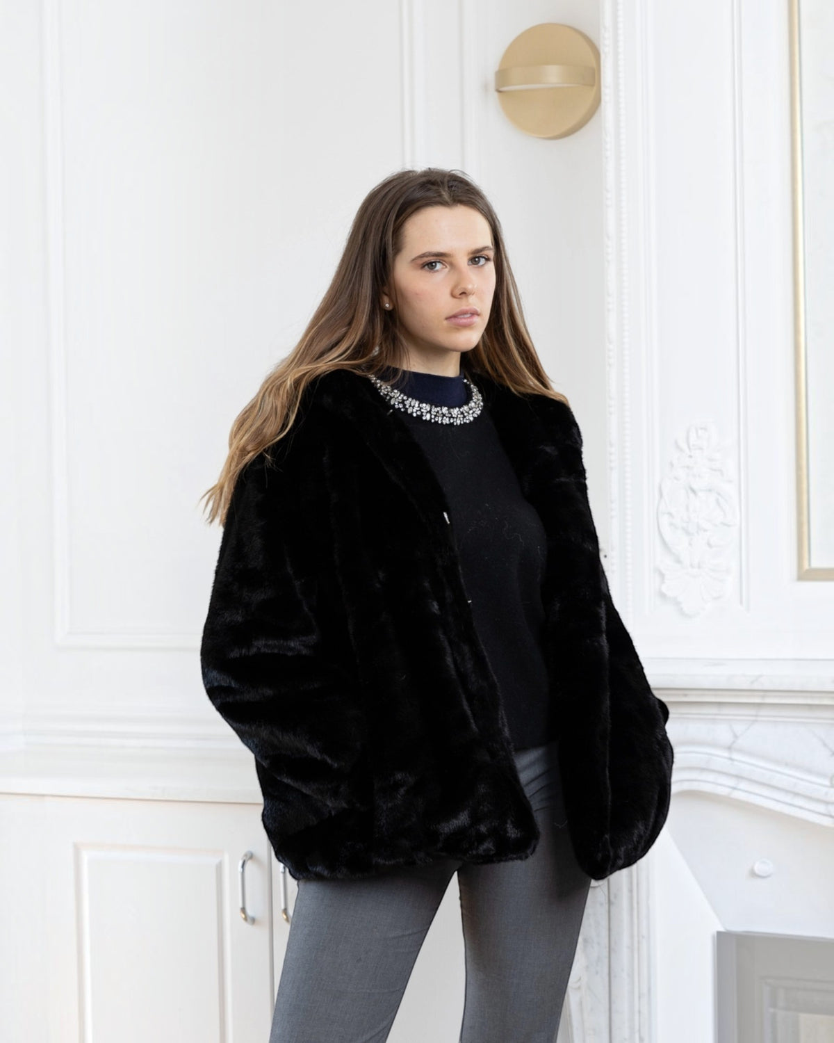 Woman wearing a black BROCK faux fur hooded coat with a velvet-touch finish in a classic interior setting.
