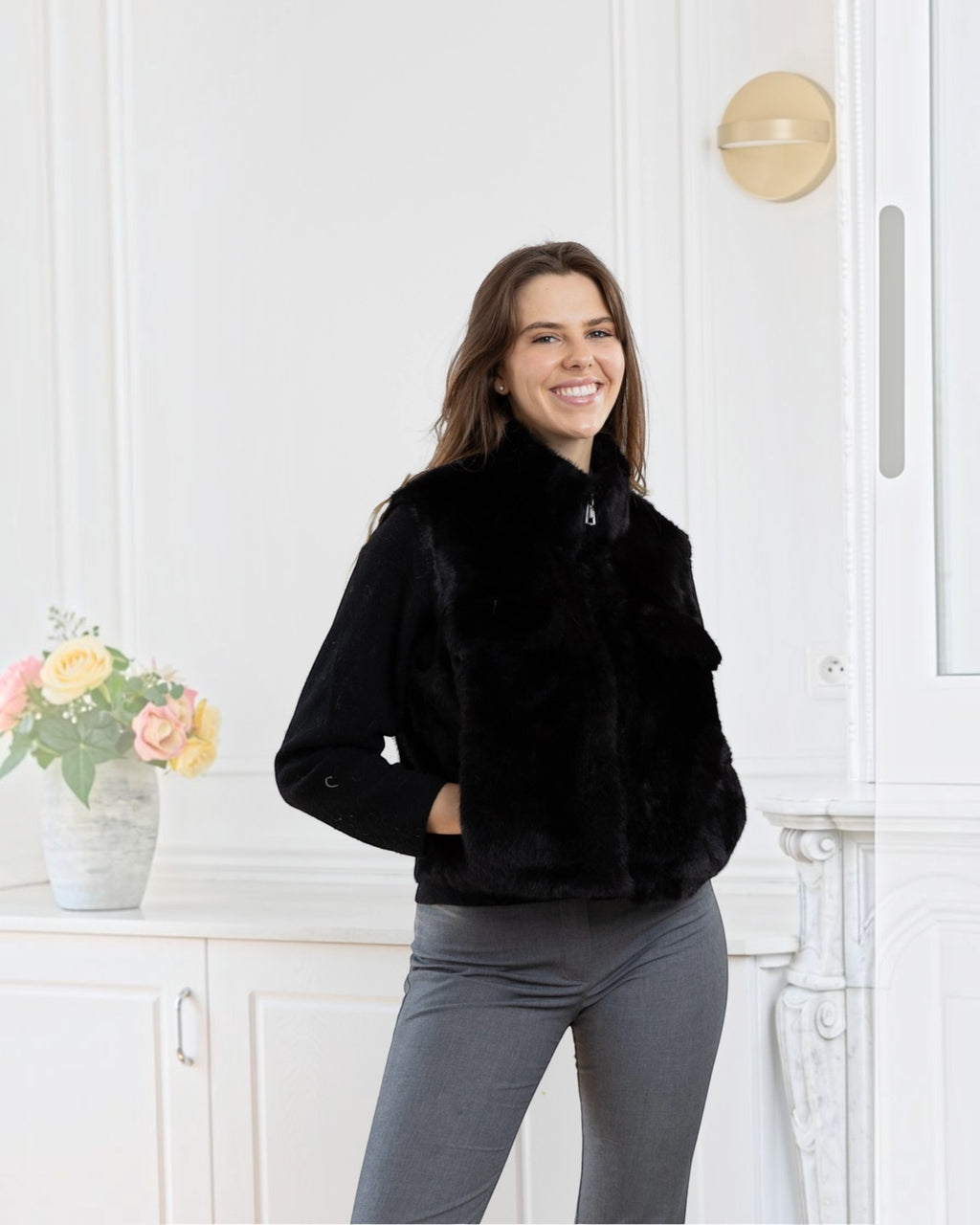 Woman wearing a black BROCK faux fur sleeveless vest with a front zipper and side pockets in a bright room.
