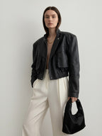 Model wearing a BROCK black faux leather cropped jacket with structured shoulders, flap pockets, and elastic hem.