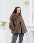 Model wearing a brown BROCK faux fur coat with a hood and velvet-touch finish in a bright, elegant interior.