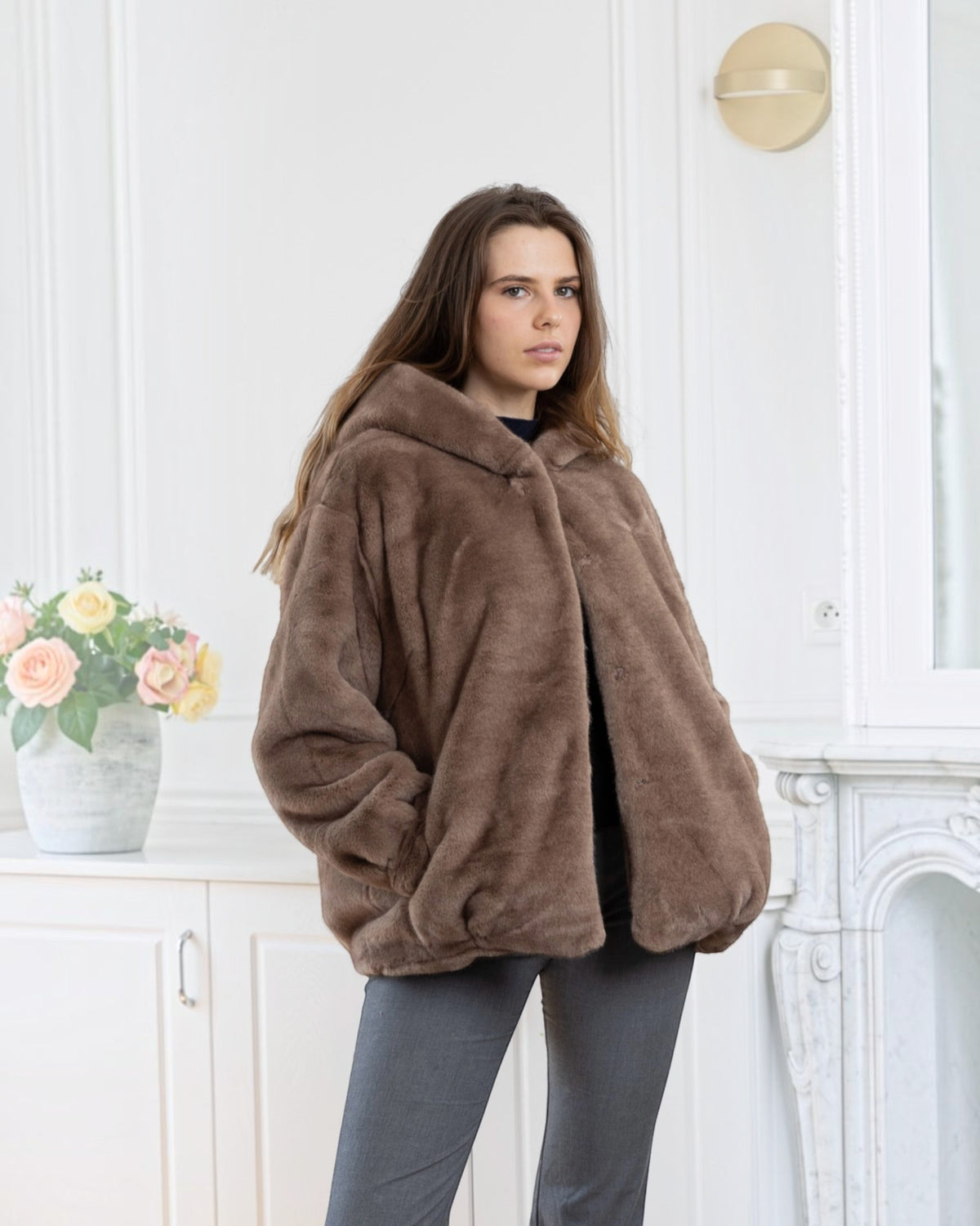 Model wearing a brown BROCK faux fur coat with a hood and velvet-touch finish in a bright, elegant interior.