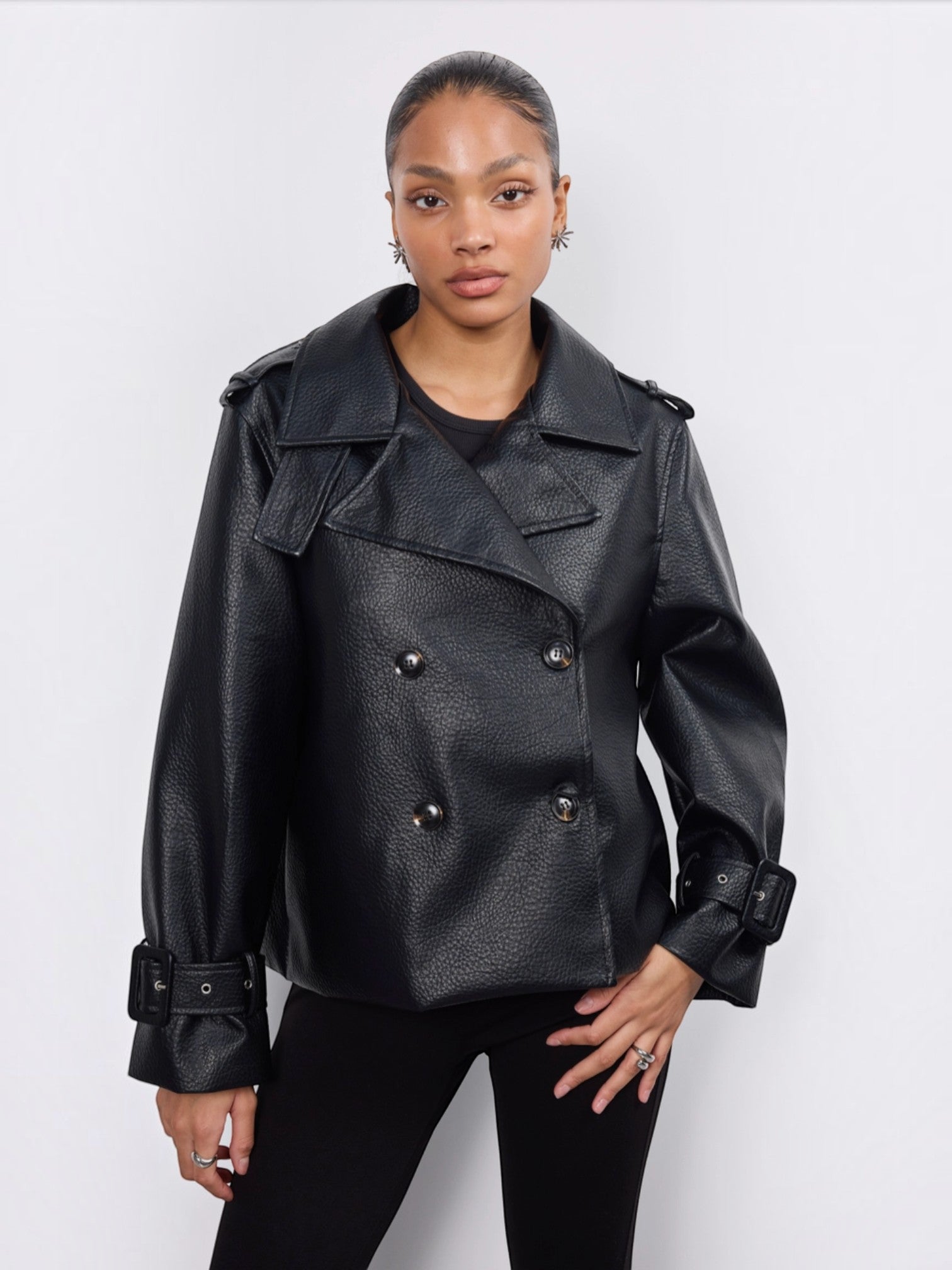 Model wearing a black BROCK Copperose double-breasted faux leather jacket with notched lapels and buckled cuffs.