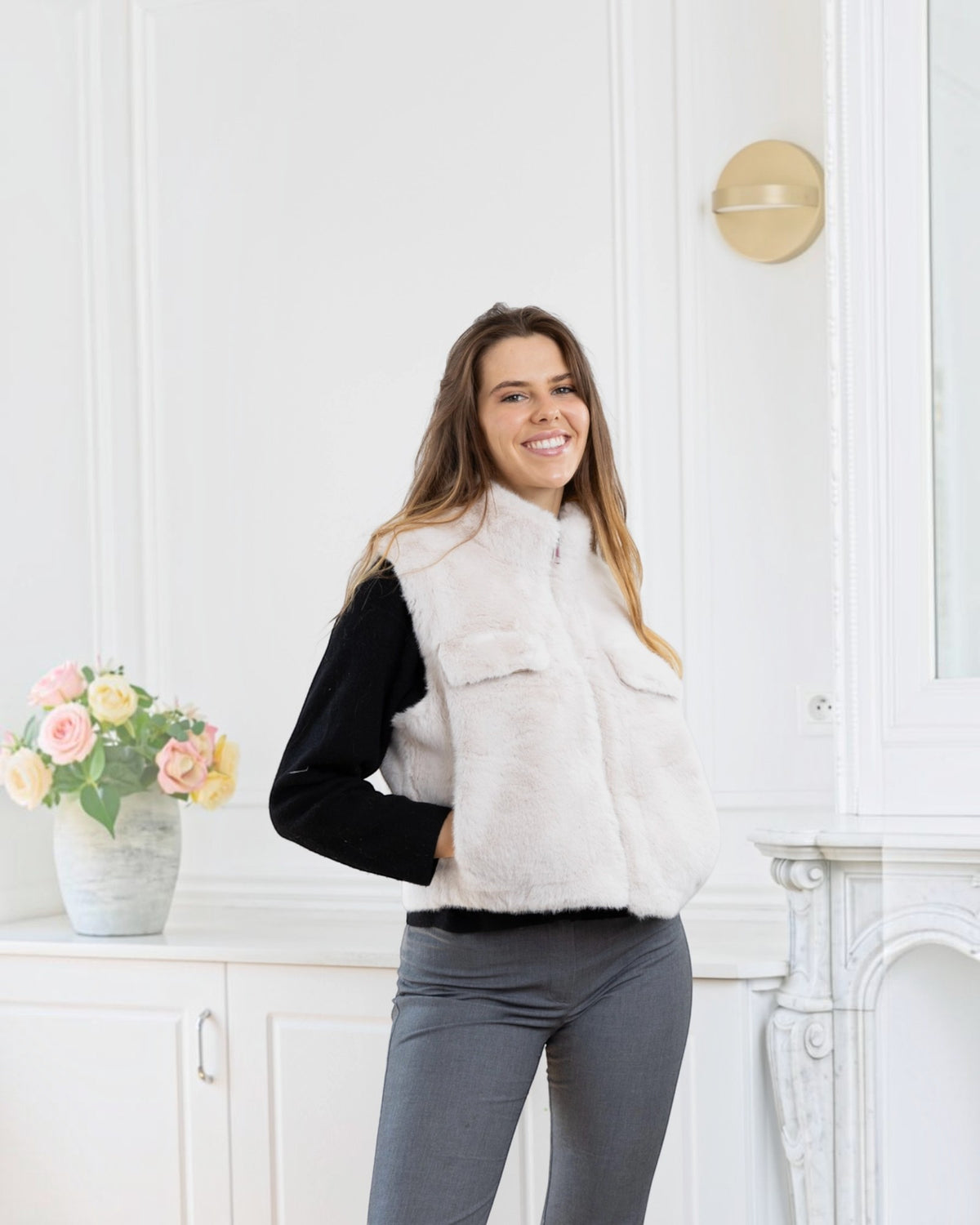 A smiling woman wearing a BROCK cream-colored faux fur sleeveless vest with a zip closure over a black sweater.