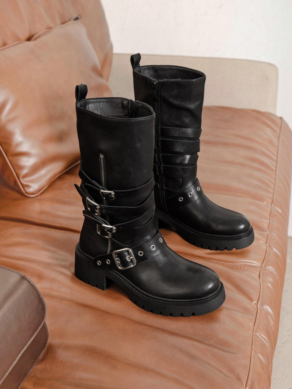 BROCK ERYNN black rock-style boots with metallic buckles, adjustable straps, and thick lug soles on a leather sofa.