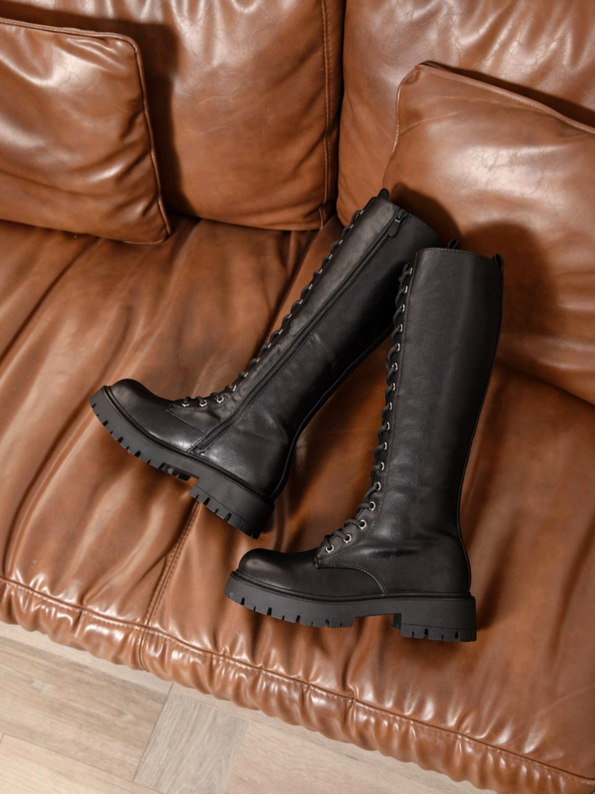 BROCK ERYNN black knee-high lace-up boots with thick lug soles and metal eyelets on a brown leather sofa