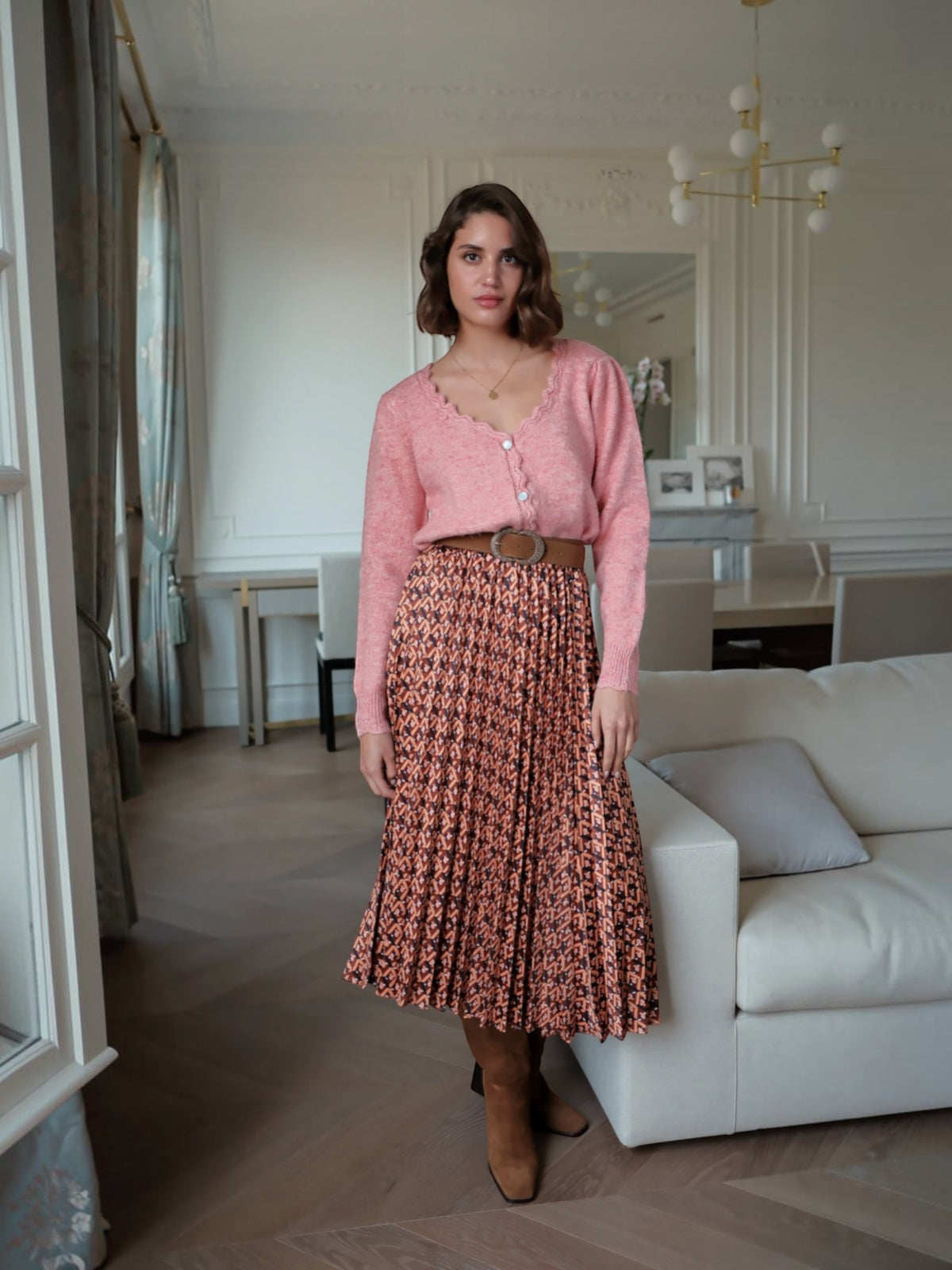 Model wearing a BROCK pleated midi skirt with geometric print, styled with a pink cardigan and brown belt
