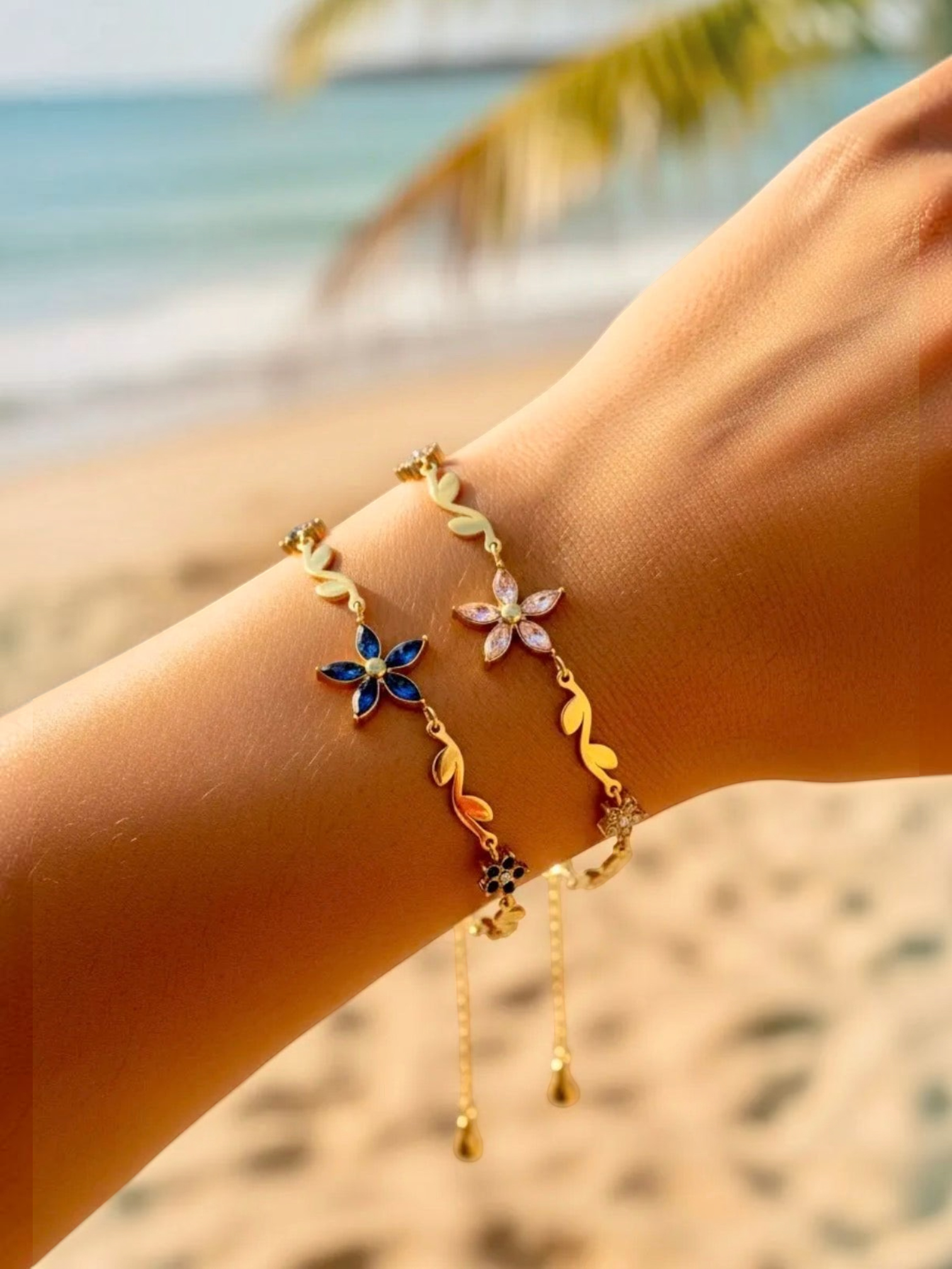BROCK gold stainless steel chain bracelet with blue and pink rhinestone flower and leaf charms on a beach background