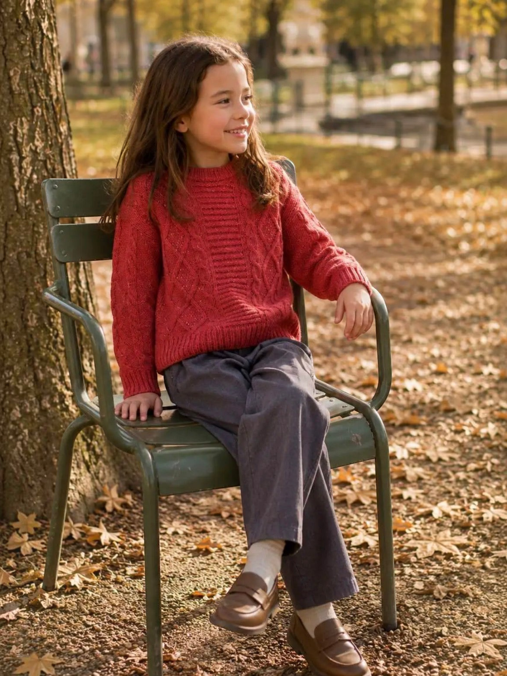 Young girl wearing a red GRASSTAR cable knit sweater and grey trousers while sitting on a park chair, BROCK brand.