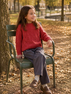 Young girl wearing a red GRASSTAR cable knit sweater and grey trousers while sitting on a park chair, BROCK brand.