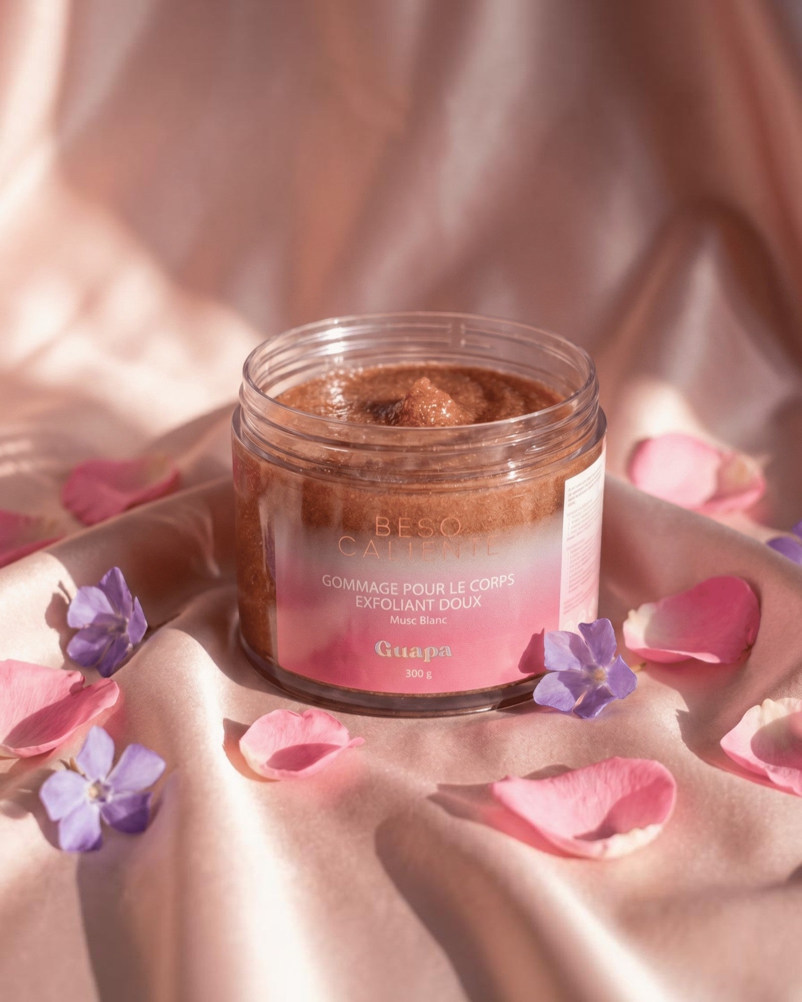 BROCK GUAPA Beso Caliente white musk body sugar scrub in a jar surrounded by pink and purple flower petals on silk
