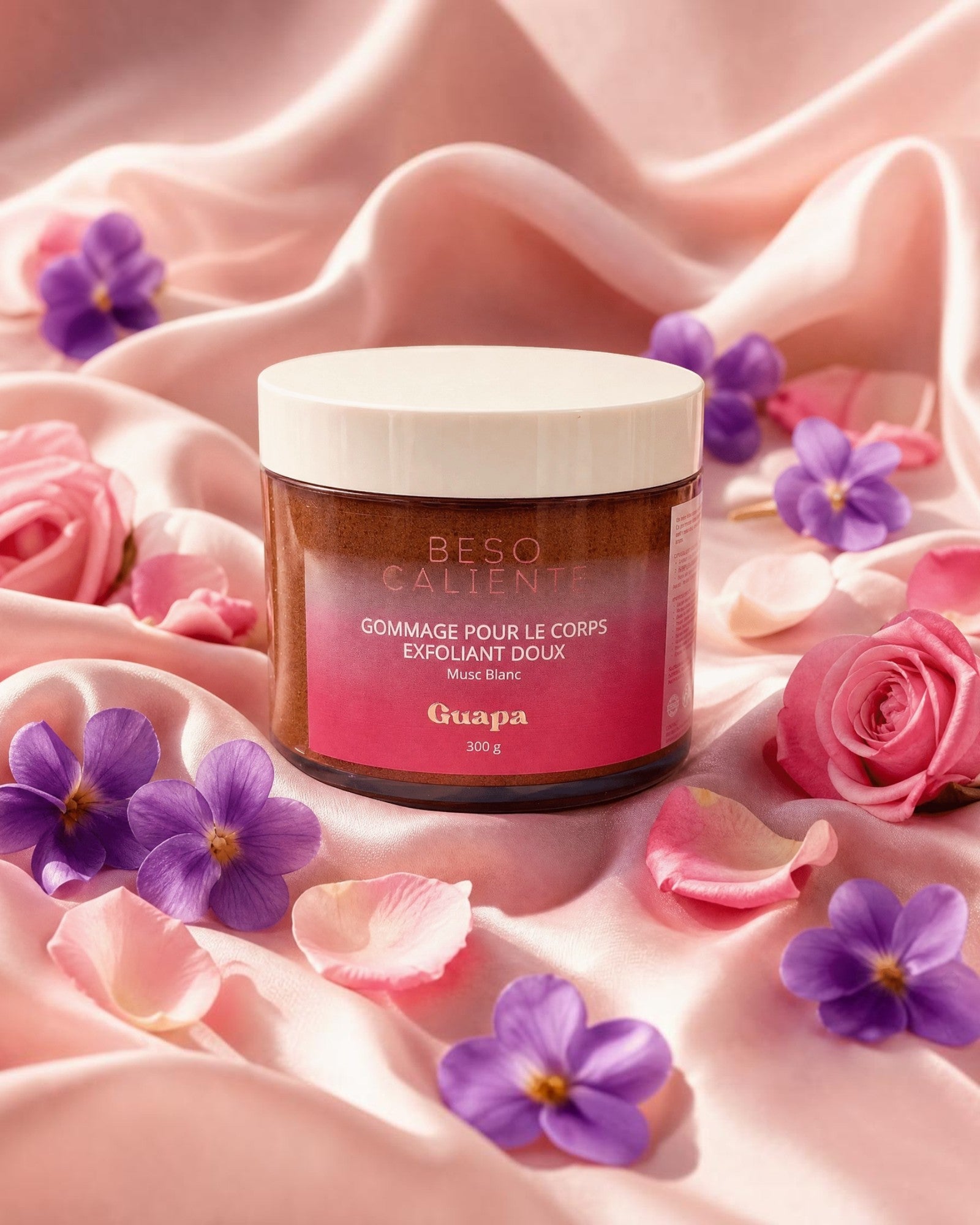 BROCK GUAPA Beso Caliente white musk body scrub in a jar surrounded by pink rose petals and purple flowers on silk