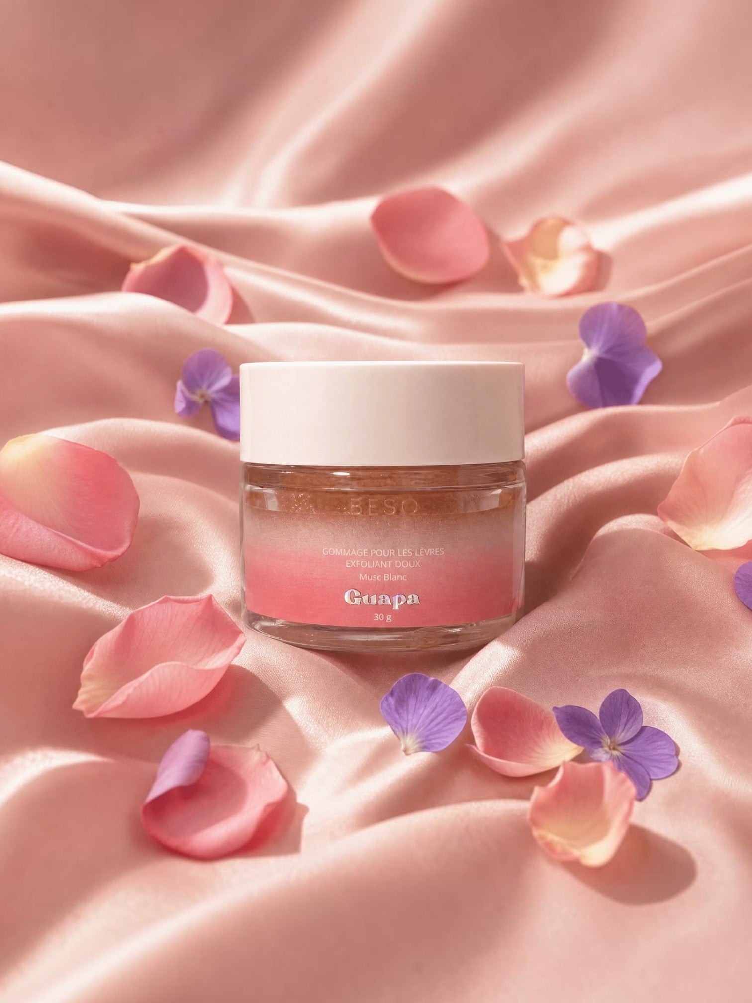 BROCK GUAPA white musk gentle lip scrub in a glass jar surrounded by pink rose petals and purple flower petals.