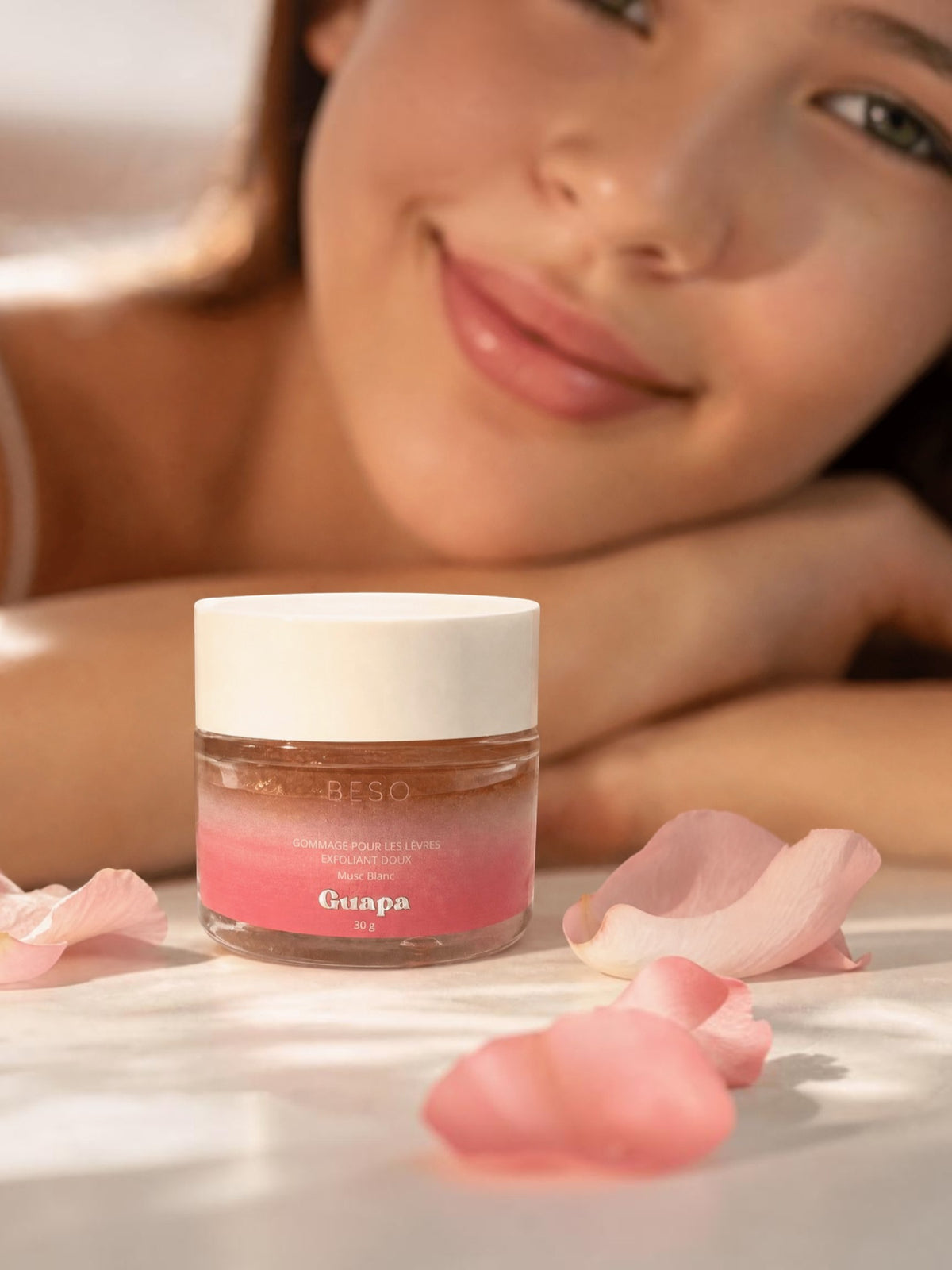 BROCK GUAPA white musk gentle lip scrub in a glass jar surrounded by pink flower petals with a smiling woman behind
