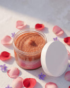 BROCK GUAPA Beso Caliente white musk sugar body scrub in an open jar surrounded by pink and purple flower petals