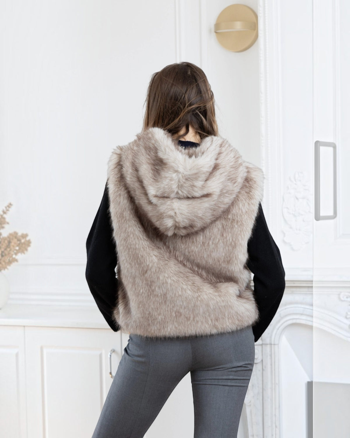 Woman wearing a BROCK LINETA beige faux fur hooded vest over a black sweater and grey trousers, back view.
