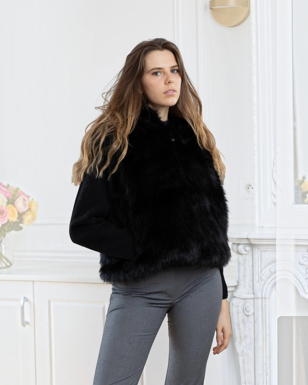 Model wearing a BROCK LINETA black faux fur sleeveless vest with a hood over a black sweater and grey trousers.