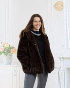 A smiling woman wearing a BROCK LINETA brown faux fur coat with a hood in a bright, elegant interior.