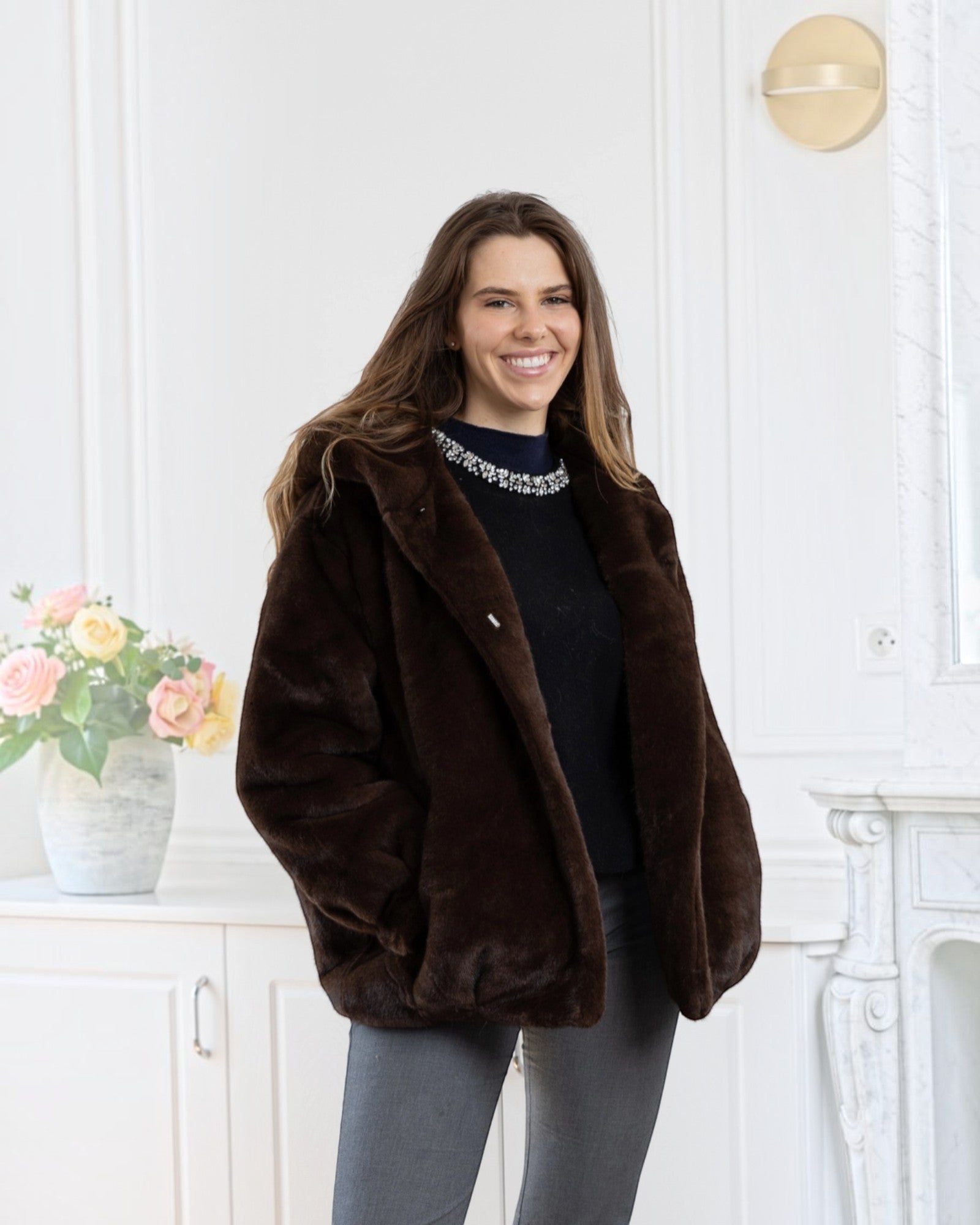 A smiling woman wearing a BROCK LINETA brown faux fur coat with a hood in a bright, elegant interior.