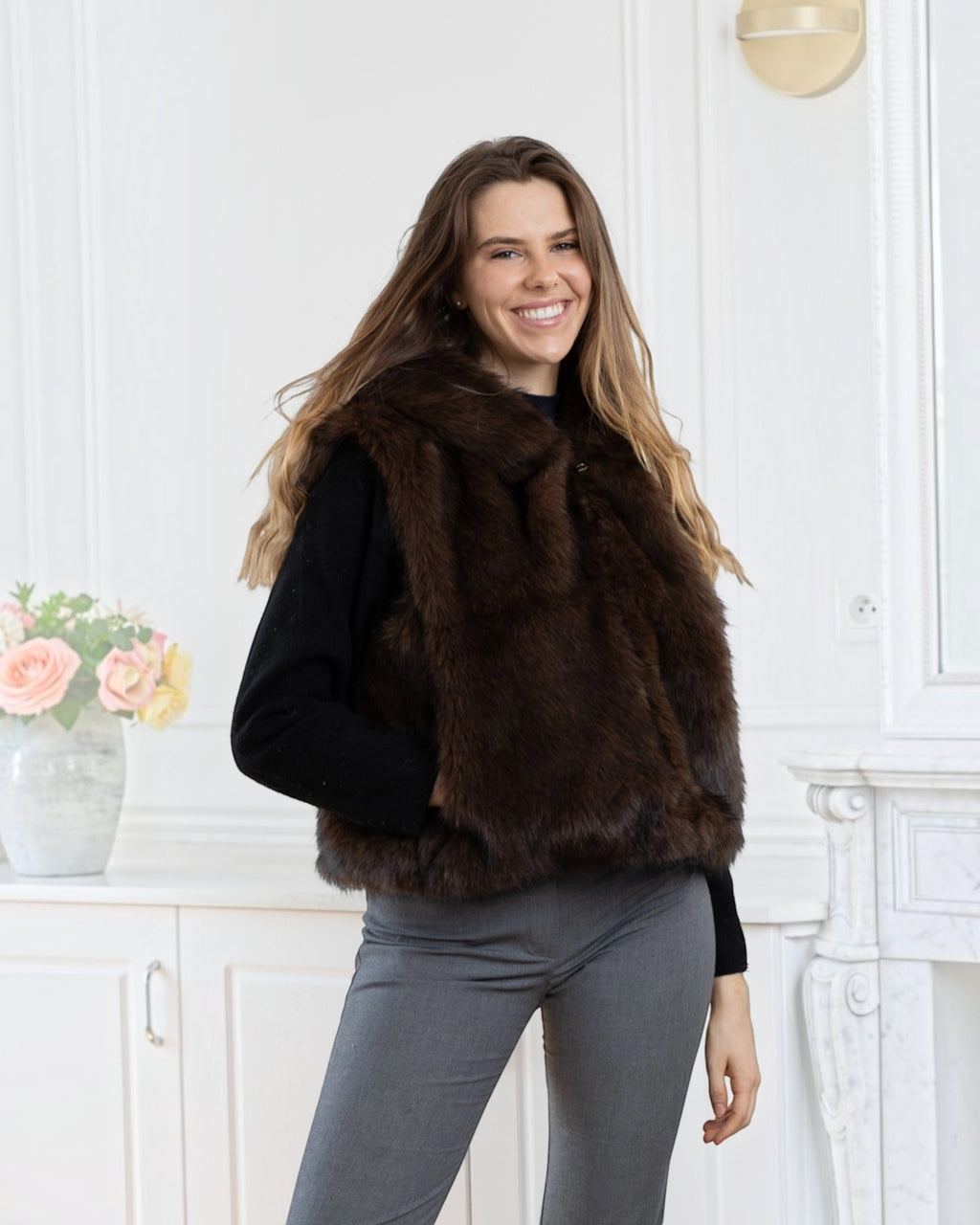 A smiling woman wearing a BROCK LINETA two-tone faux fur hooded vest over a black sweater and grey trousers.