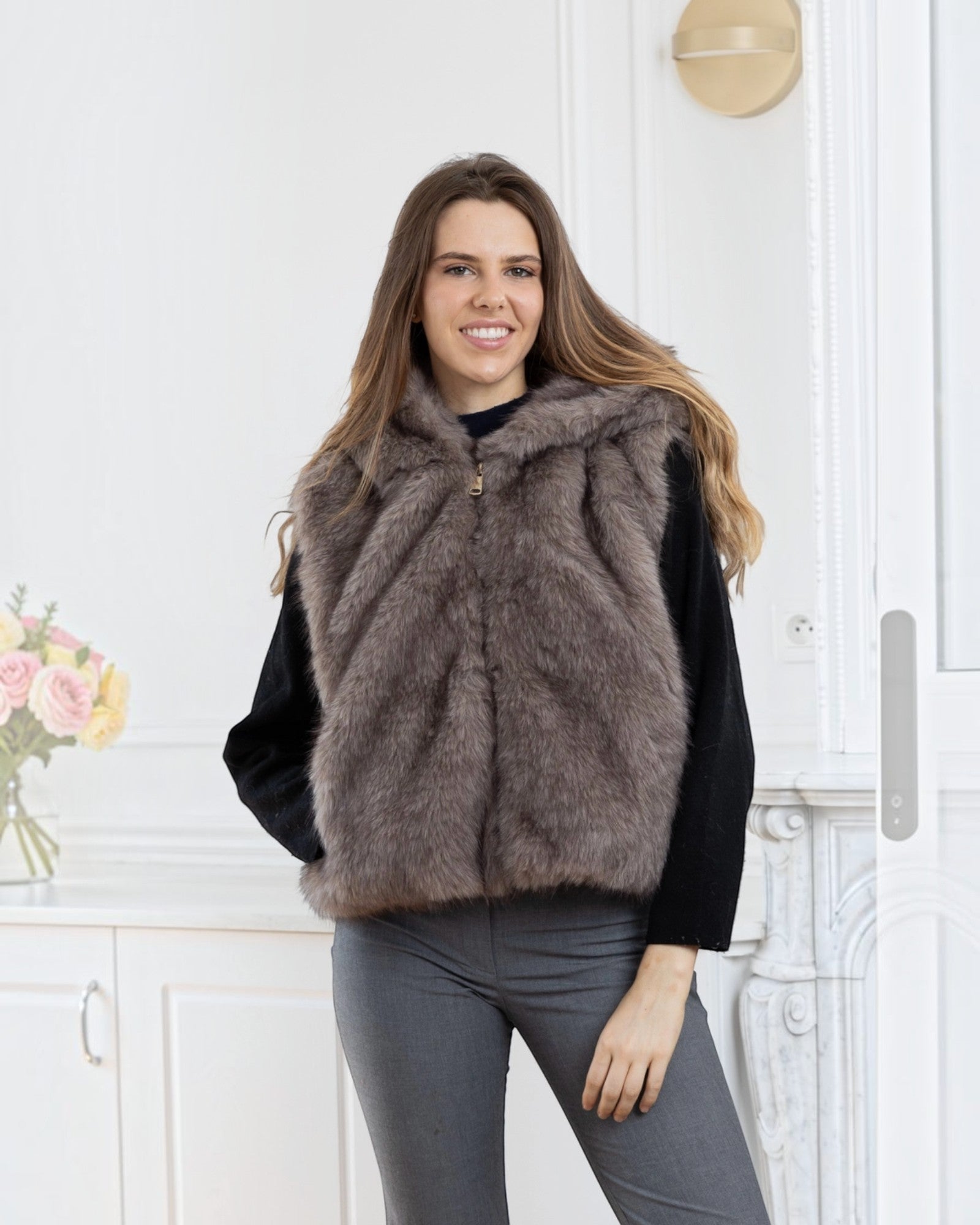 BROCK LINETA sleeveless two-tone faux fur vest with hood, worn by a smiling woman in a bright interior.