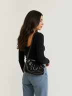 Model wearing a BROCK LOUISA LEE black metallic leather croissant crossbody bag with a ruched design