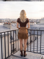 BROCK MOOYA brown faux suede mini skirt with fringe hem and matching belt, worn by a blonde woman on a city balcony.