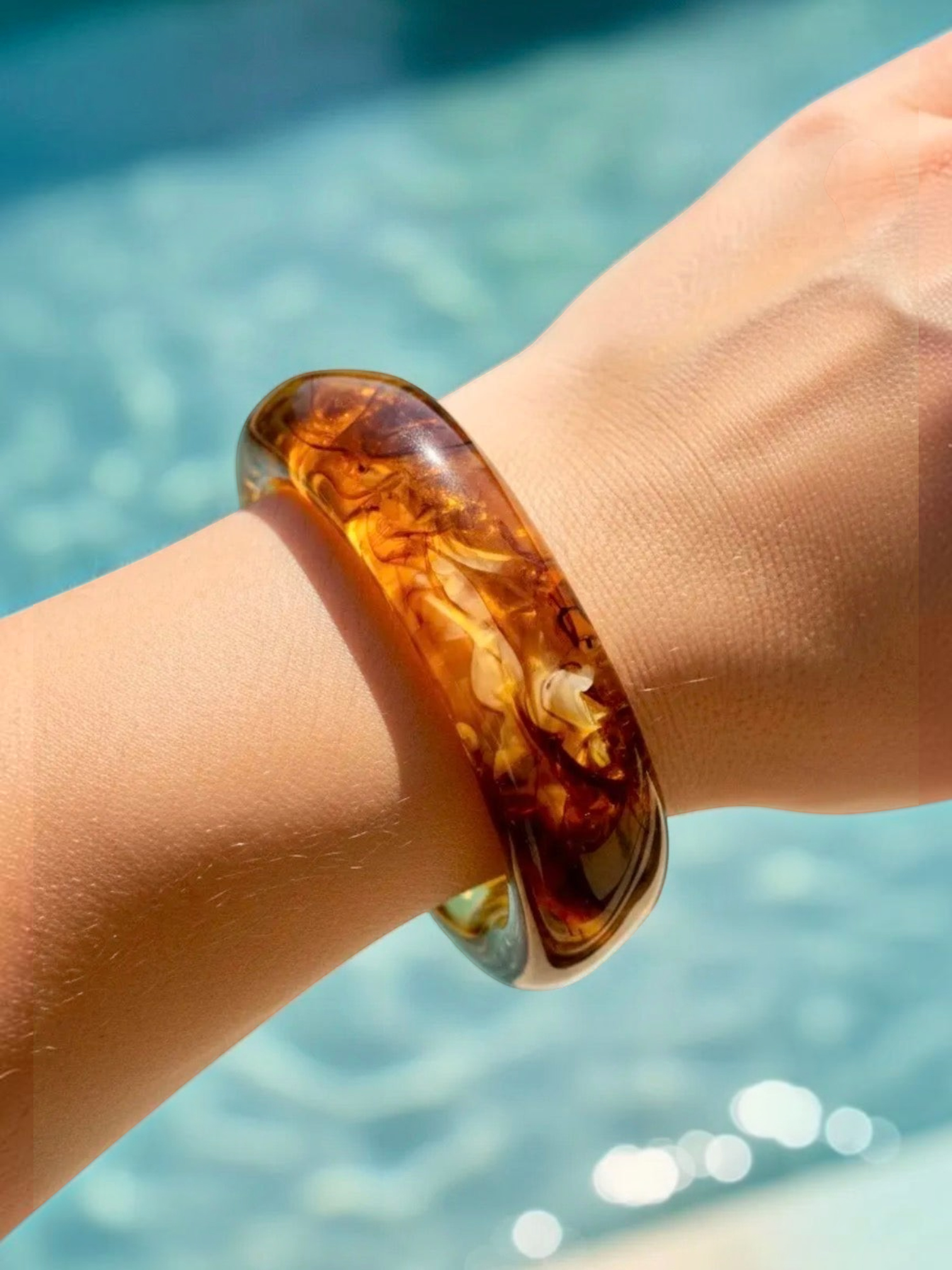 BROCK ROSA amber resin bangle bracelet with golden inclusions worn on a wrist against a blurred pool background