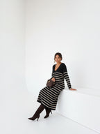 Model wearing a BROCK long-sleeved striped maxi dress with a V-neck, styled with dark boots in a minimalist studio.
