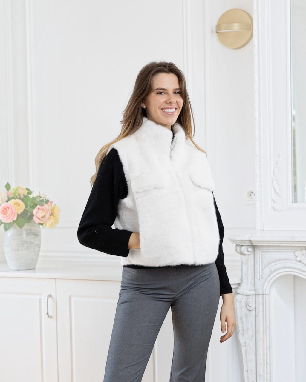Model wearing a white BROCK faux fur sleeveless vest with a zip closure and side pockets in a bright room.