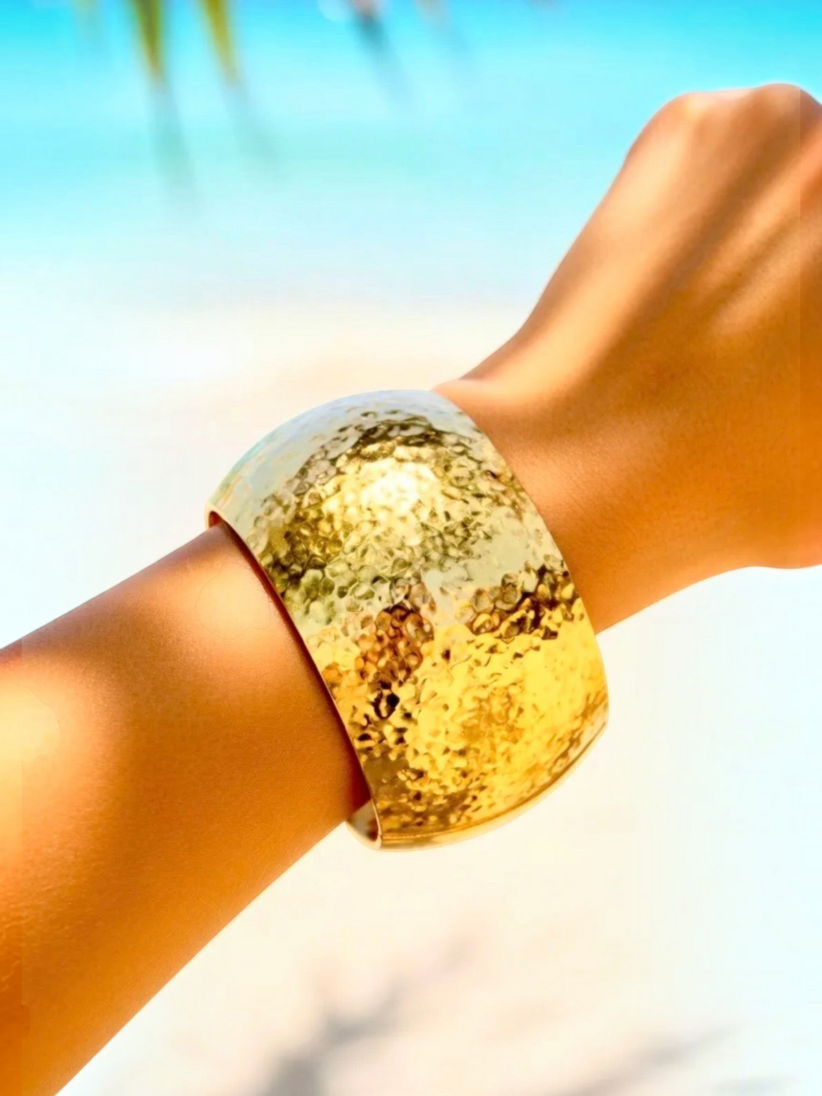 BROCK wide gold hammered brass cuff bracelet worn on a wrist against a blurred beach background