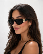 Lunettes de soleil ÉLINE L'ATELIER noires avec détails dorés sur les branches, monture épaisse et verres teintés.