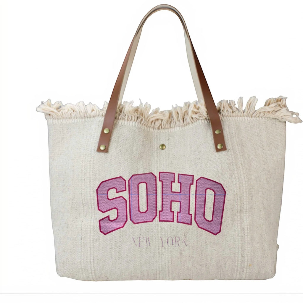 Cabas en coton beige avec anses en cuir, inscription SOHO rose et finitions à franges, style décontracté.